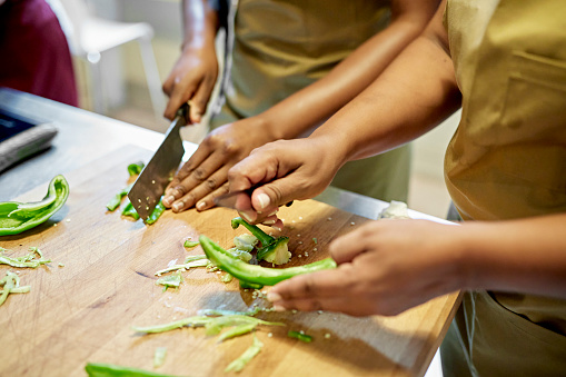 Close-up of African and Colombian women in aprons standing at cutting board slicing green bell peppers.